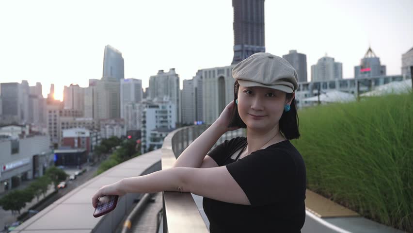 A woman in a light beret and black top poses on a curved rooftop with a city skyline at dusk in the background.