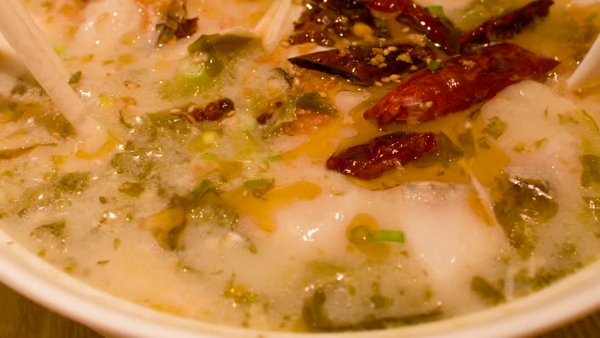 Macro shot of chopsticks and spoon stirring spicy fish noodle soup in a ceramic bowl.
