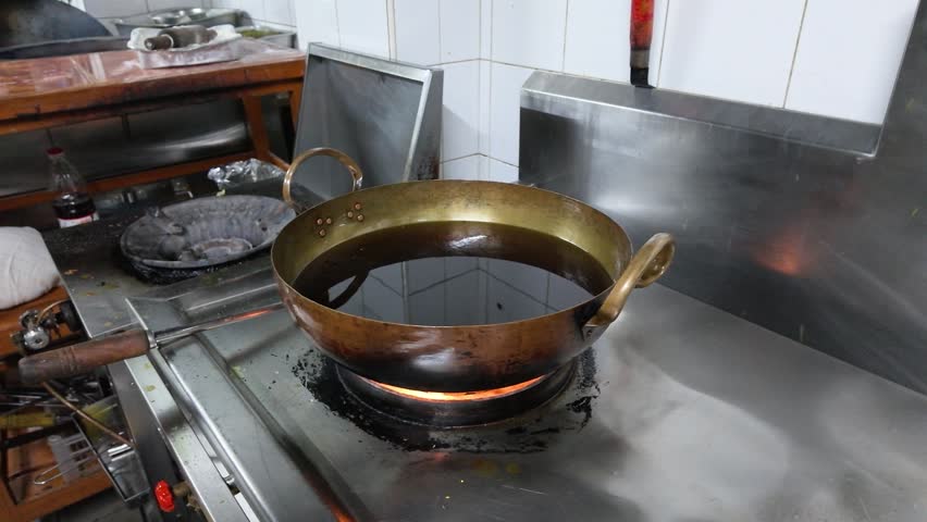 Static medium shot of a brass wok heating oil over an open gas flame.