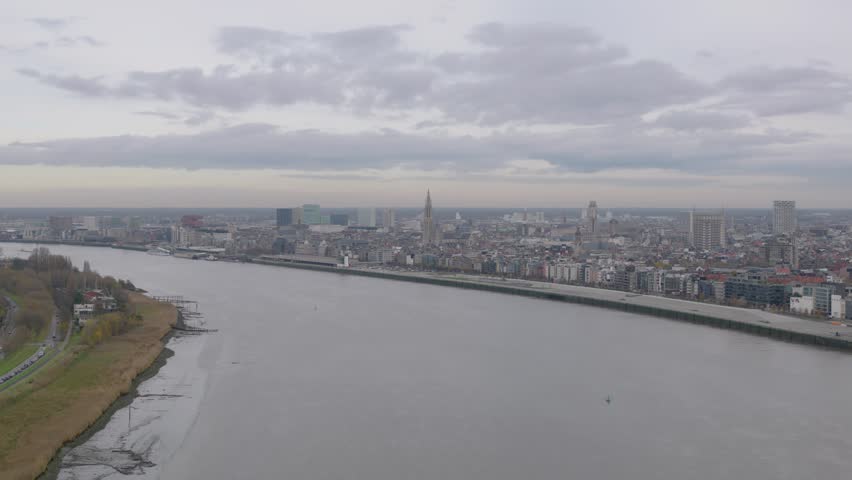 Drone footage showing a wide view of Antwerp with the Scheldt River, the quays, and the city skyline under a cloudy sky.