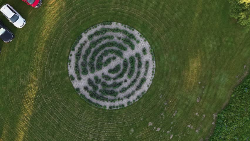 Drone video captures a symmetrical labyrinth garden surrounded by manicured grass and several parked cars, filmed in soft late afternoon sunlight with crisp, vibrant colors from above.