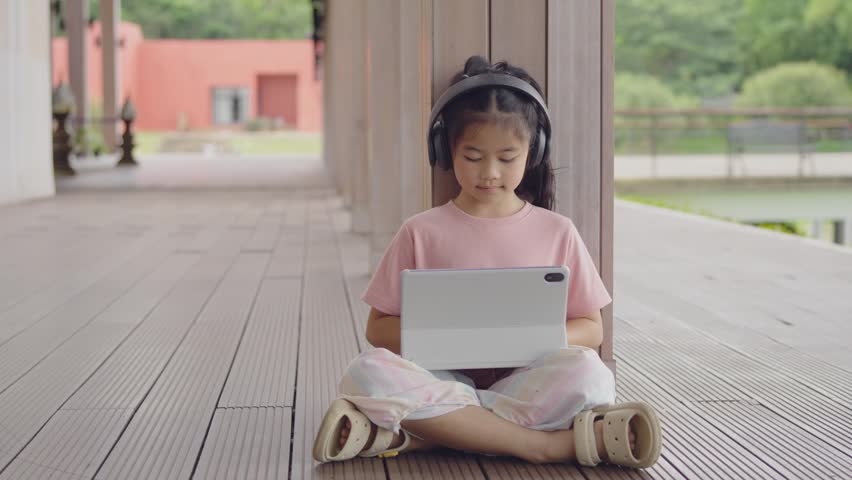 Young girl sitting cross-legged outdoors on wooden decking, wearing large headphones and pink t-shirt, using a tablet device on bright day