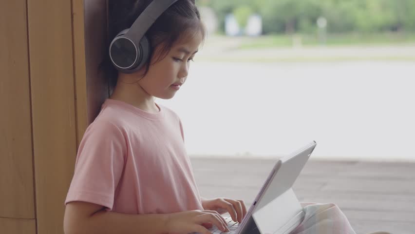 Young Girl Wearing headphones and using tablet device while leaning against wooden wall outdoors on a bright day