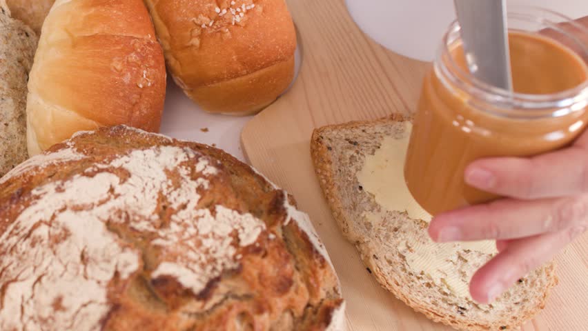 Close-up shot of hands spreading peanut butter over buttered multigrain bread on wooden board.