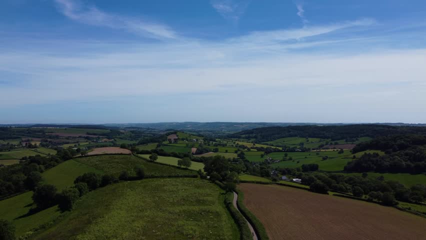 Green farmland, hedgerows and distant hills form a wide rural landscape under a clear blue sky.