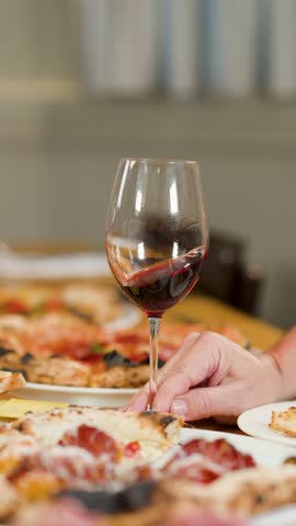 Close-up of a hand holding red wine beside pepperoni pizza on a restaurant table.