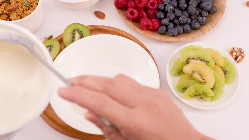 Overhead shot of hand scooping thick yogurt into bowl surrounded by fresh fruit.