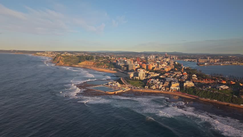 Aerial panorama of Newcastle city and ocean baths beside rolling surf at sunrise, drone high overview approach