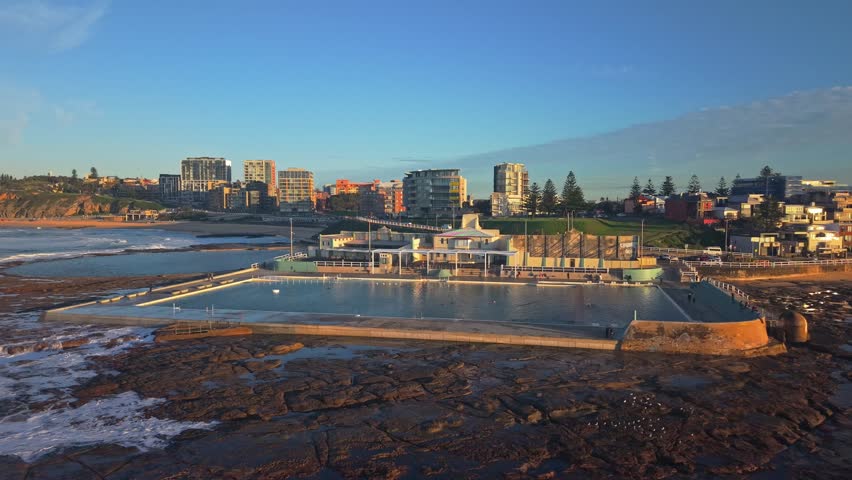 Newcastle Ocean Baths glow in sunrise light beside crashing waves and city skyline, drone panoramc pullback as waves crash on rocks