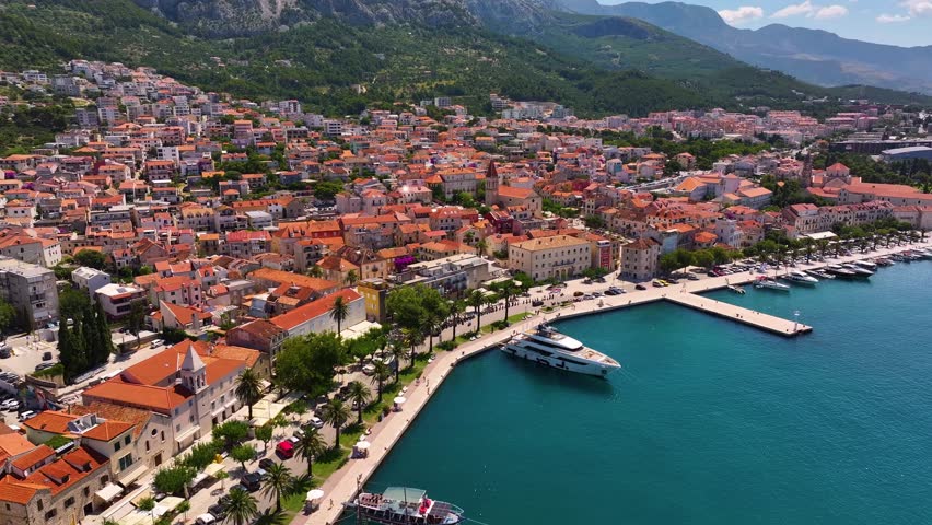 Drone shows Makarska Croatia with buildings along coastline. Boats docked in harbor. Mountains rise in background. Daytime clear weather.