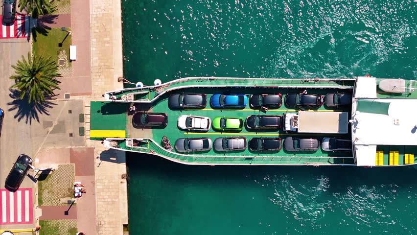 Cars are lined up and loading onto a ferry at the harbor. The ferry is docked near the beach. People are waiting on the dock.
