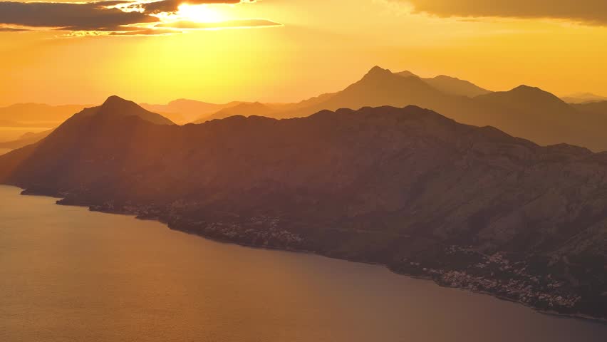 Sunlight reflects off water as the sun sets behind mountains. The landscape includes hills and a coastal area. Color changes in the sky are visible.
