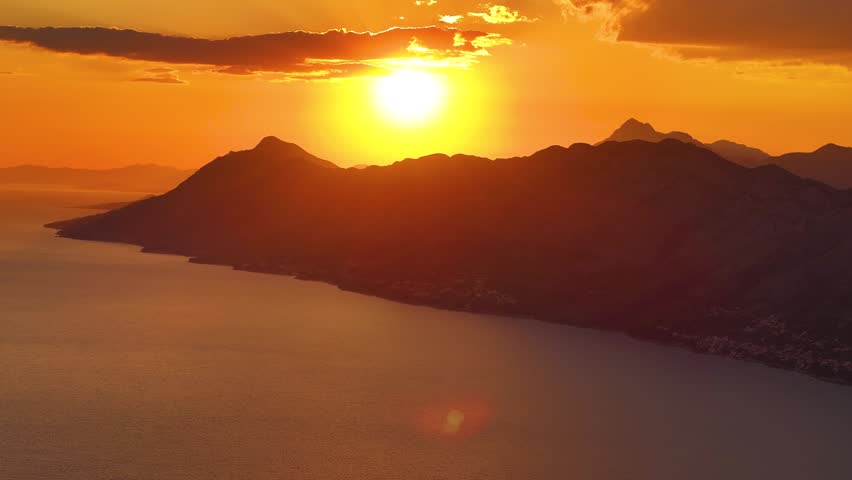 Sun sets over mountains and water. Light reflects on the surface. Silhouettes of hills are visible against the horizon. Scene shows natural landscape.