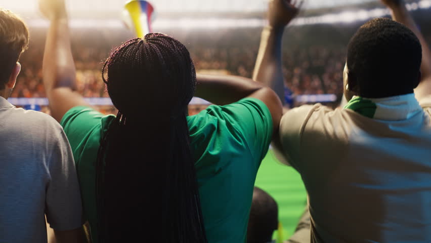 Soccer supporters cheer in packed stadium stands during a football match, creating loud crowd energy and team support, with raised hands visible across the sports arena.