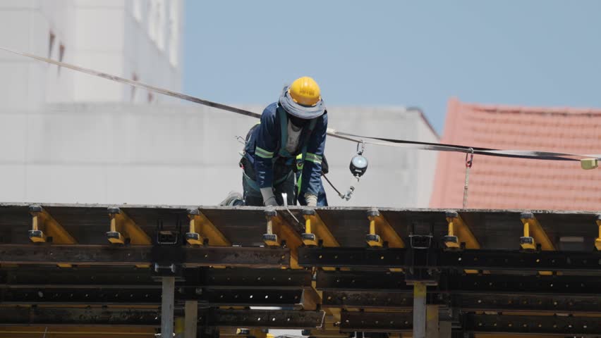 A worker is seen on a construction site. He is installing materials on a building. The sun shines brightly above him in the clear sky. Work safety gear is worn