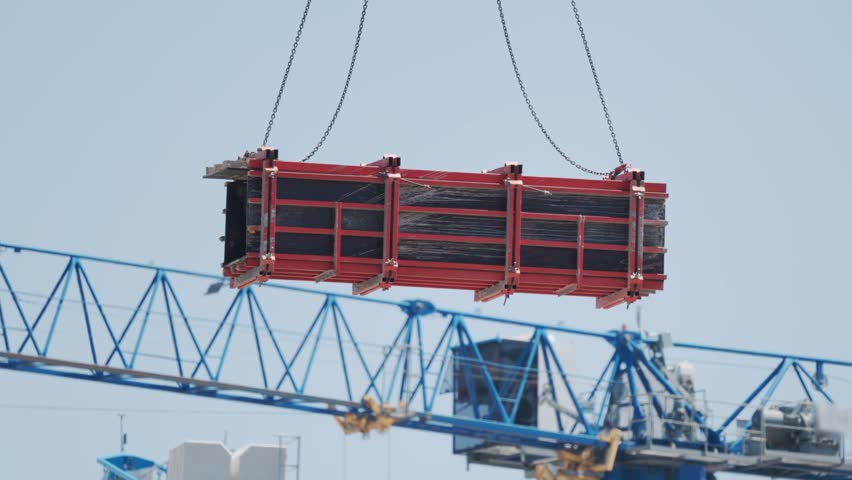 Workers operate a crane to lift a large, red container at a construction site. The sky is clear, and other cranes are visible in the background