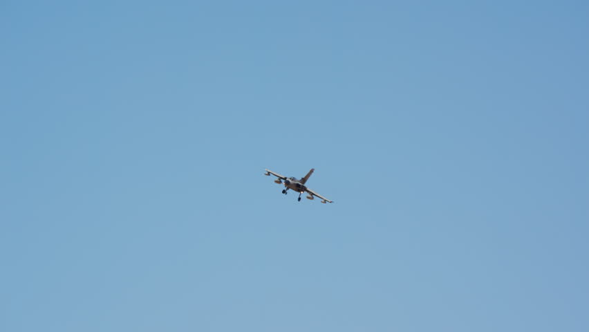 Military fighter jet banking on approach with gear down seen from below, combat aircraft turning against blue sky, warplane descending toward the runway to land