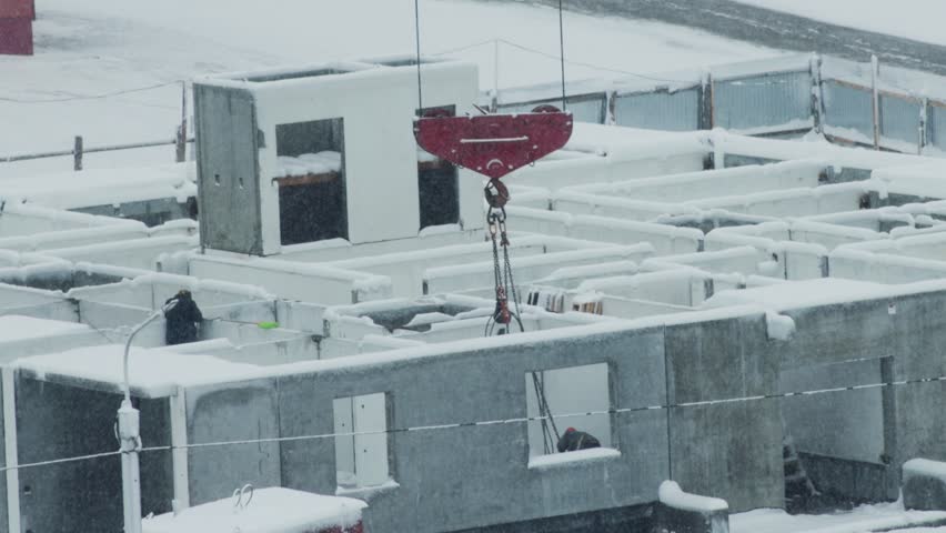 Overhead view of building construction site, covered in deep snow, heavy flakes falling. Red crane hook lowers large white pre-fabricated concrete wall section into place. One person, bundled in winte