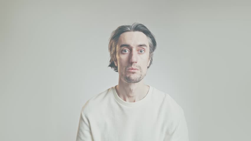 Surprised adult man making shocked and curious facial expressions in minimalist studio, soft lighting, light gray background, centered portrait with copy space