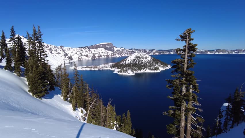 A landscape view of Crater Lake National Park with trees, blue sky and Wizard Island. The snowy is covering the area under clear sky in Oregon