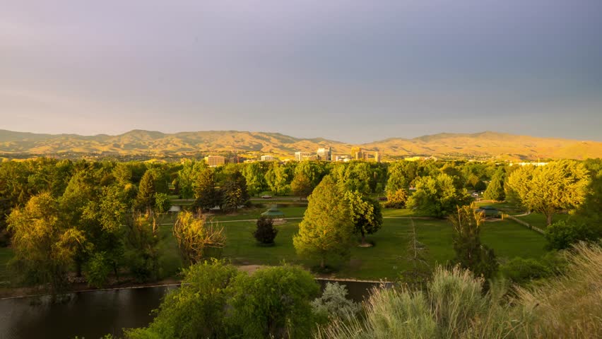 Aerial timelapse of Ann Morrison Park in Boise Idaho featuring lush trees a winding river and a distant city skyline under a vibrant shifting sunset sky with beautiful evening light