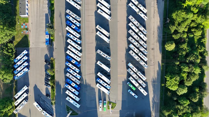 Aerial view of a bus fleet parked in a lot, showcasing a continuous arrangement of buses with surrounding trees and buildings, highlighting the organized layout and spatial relationships