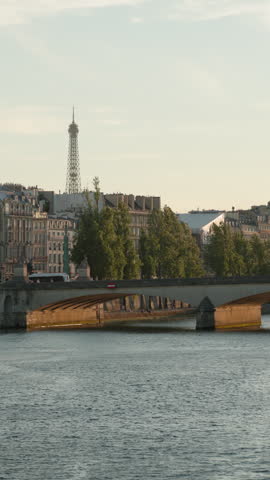Vertical view of a classic stone bridge arching over the Seine River in Paris, with historic Parisian buildings and the iconic Eiffel Tower rising elegantly into the soft afternoon sky.