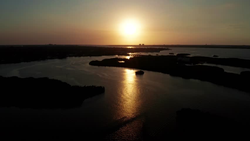 Sunset glows across an estuary as low forested islands cast dark shapes and rippled water reflects the sun from a high drone view.