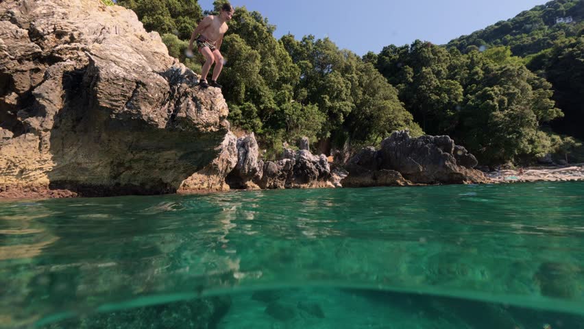 Split shot of a happy teen boy jumping and diving into the clear sea water with splash, over under half underwater view near rocky shore.