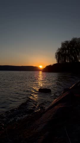 Scenic landscape view of a peaceful lake at dusk with the sun dipping below the distant horizon hills.