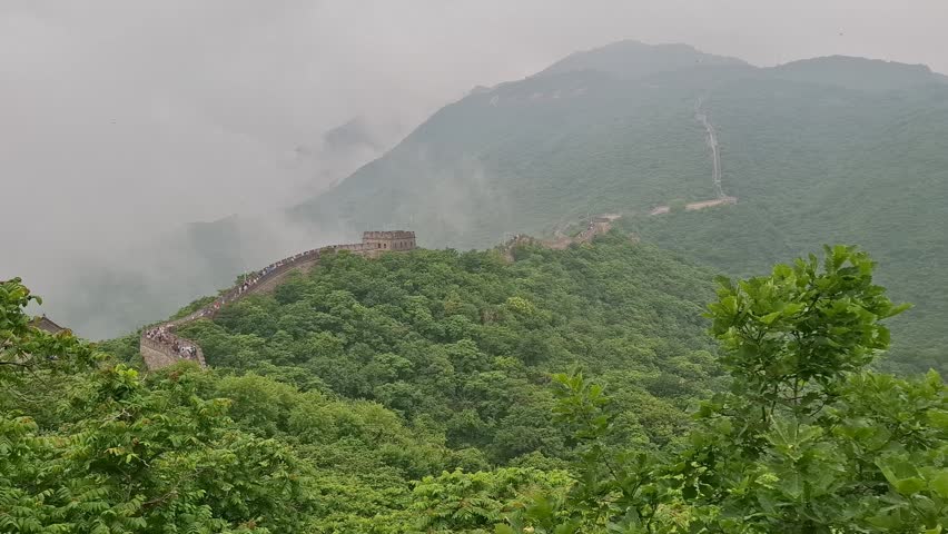 Scenic view Great Wall of China in Mutianyu, near Beijing, snaking across green mountain ridges partially covered in heavy low fog and mist. Ancient stone fortifications. Famous Asian travel  landmark