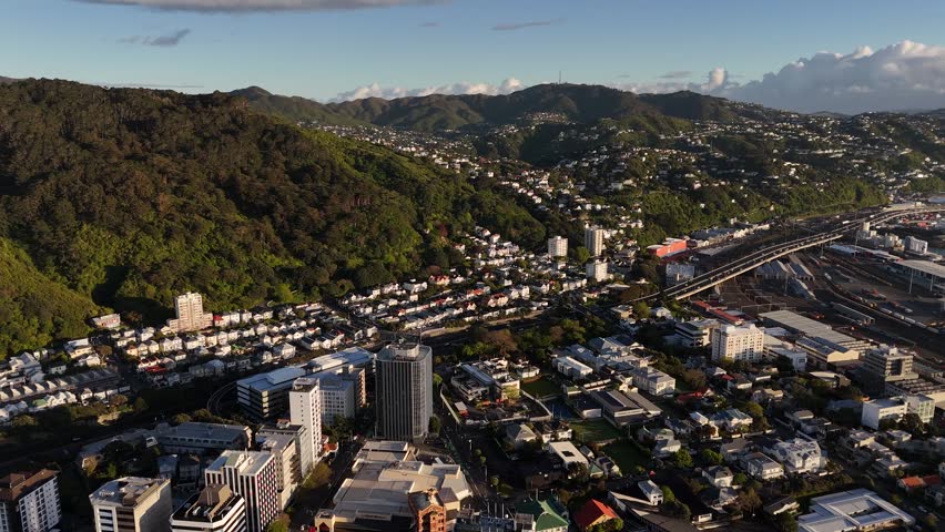 Hillside homes and downtown towers spread across green hills above sprawling rail yard as warm golden sunrise light bathes Wellington’s busy city center. New Zealand. Aerial drone view.