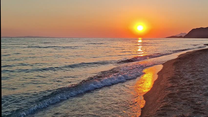 Beautiful golden sunset over Vrachos beach in Greece, sea with calm waves crashing on the sandy shore.