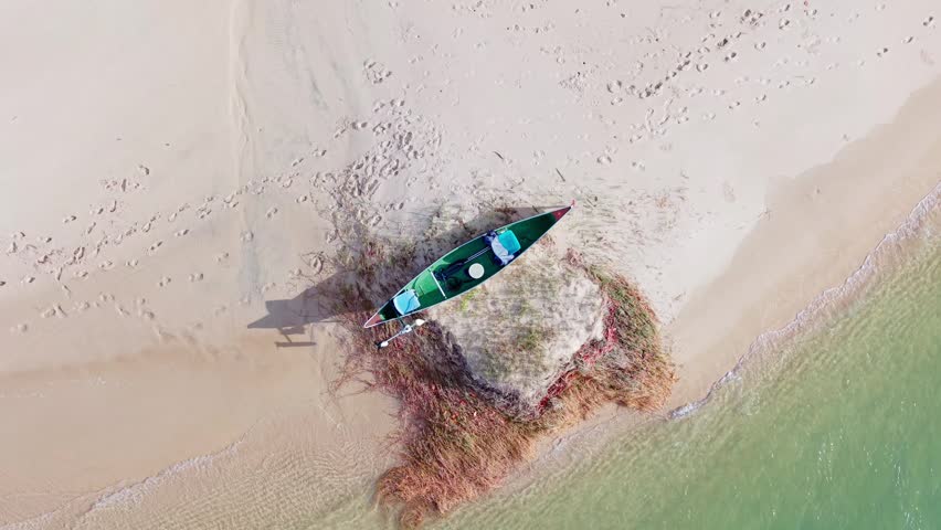 Top down drone aerial of a small canoe resting on a sandy shoreline beside shallow turquoise water at the Noosa River estuary in Queensland Australia.