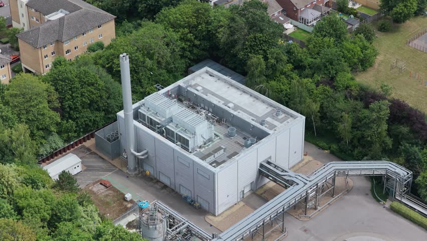 An aerial perspective of a contemporary energy center, featuring a tall chimney and complex pipe systems, situated among lush green trees and residential buildings.