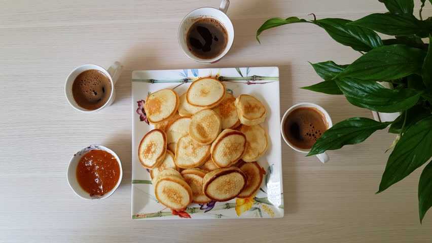 Top view of multiple generations of family hands taking small pancakes from a white plate and dipping them in apricot jam. Breakfast scene with three cups of coffee.