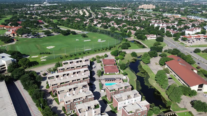 Multifamily residences in Irving, Texas overlook manicured fairways winding Elm Fork Trinity River waterways. Rooftop terraces swimming pools sit beside dense summer foliage near concrete spillway. US
