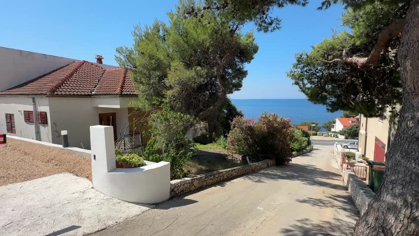 A road slopes down towards the sea in a sunny town. There are houses on both sides of the road, and trees provide some shade. The blue water is visible in the distance.