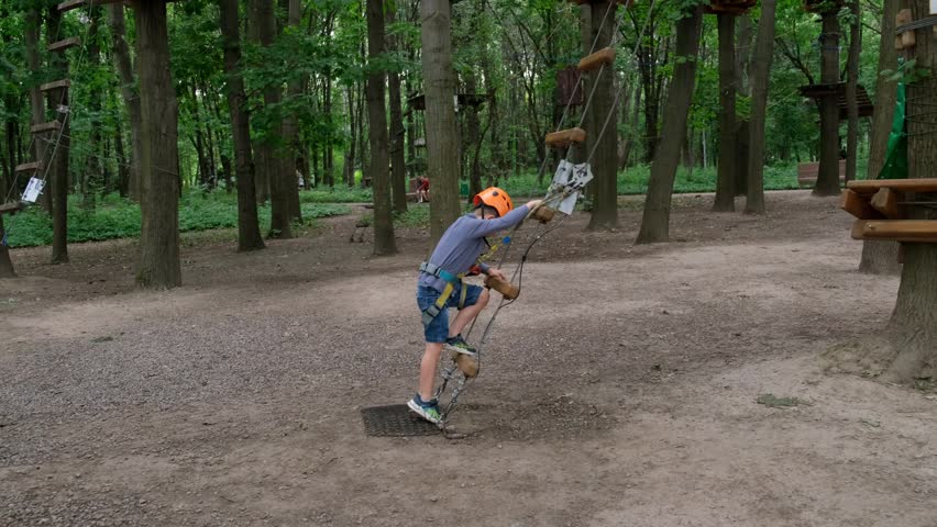 A brave young boy wearing an orange safety helmet and harness climbs up a rope ladder to start an obstacle course in a forest adventure park.