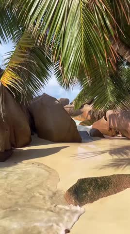 Gentle sea waves crashing on a tropical sandy beach with large granite rocks and palm trees