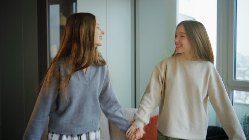 Two teenage friends hold hands and joyfully fall onto a sofa in a cozy apartment, continuing their cheerful conversation in a warm and relaxed atmosphere