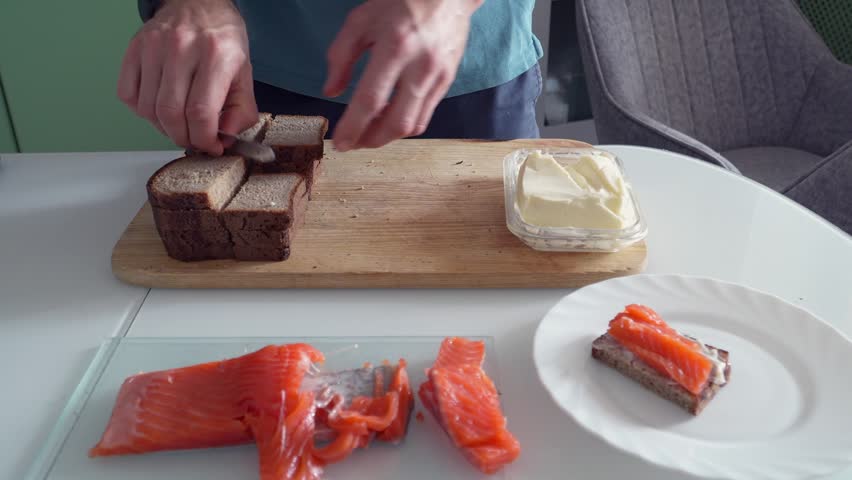 man making sandwiches with butter and salmon front view, kitchen food preparation process with slicing bread and spreading butter, preparing snack meal, close-up action assembling fish sandwich