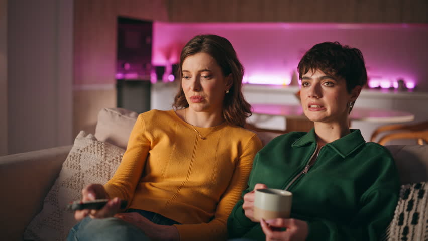 Sisters choosing tv movie switching channels sitting together cozy living room couch. Relaxed two women watching television enjoying domestic evening atmosphere. Happy friends relaxing on home sofa.