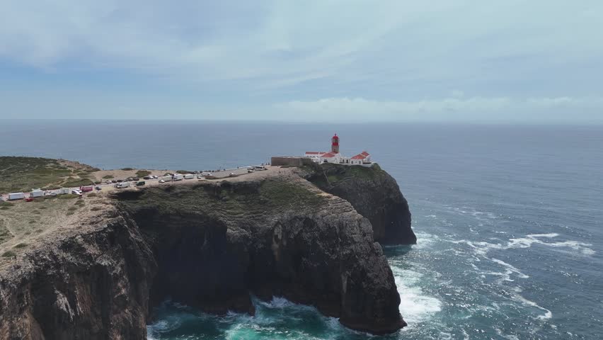 Aerial drone shot panning right and tracking rugged cliffs of Cabo de São Vicente, near Sagres, Portugal. The historic lighthouse overlooks the ocean, with waves below.