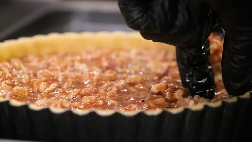 Close-up of a pastry chef wearing a black nitrile glove carefully arranging walnut halves in a rich caramel-filled tart shell. The footage highlights the gooey texture of the caramel and the crunchy w