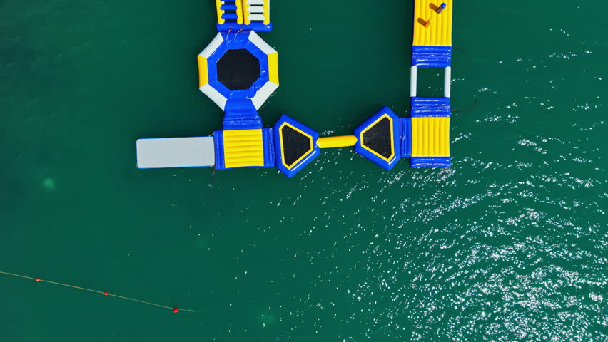 A colorful inflatable water park sits on the surface of blue water. People jump and play on the floating structures while enjoying their time outdoors in the sun.