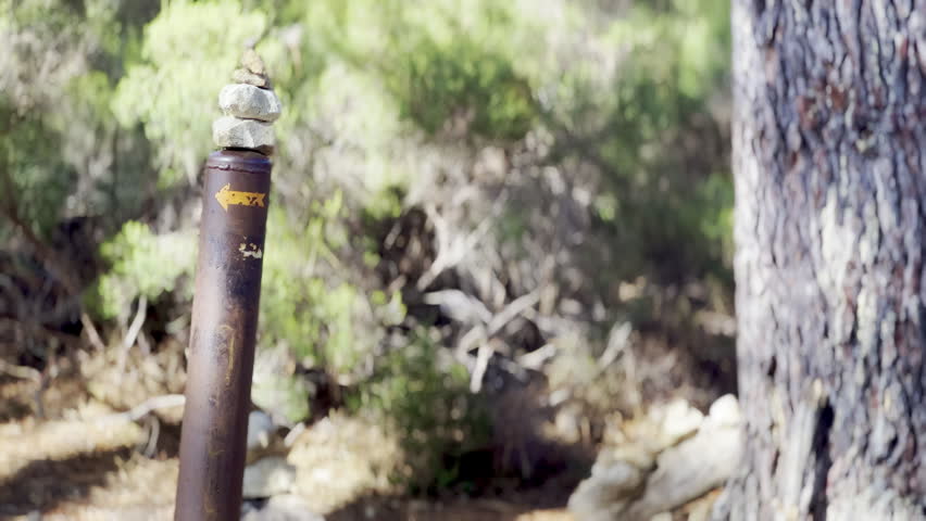 Small balanced stone cairn on top of a rusty hiking trail marker post with a painted yellow arrow beside a pine tree in the Luberon forest, Provence, France