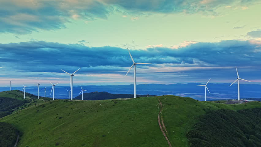 Wind turbines spin on rolling green hills in a rural area. The sky is filled with clouds that change colors as the sun sets. The wind moves through the blades, creating clean energy.