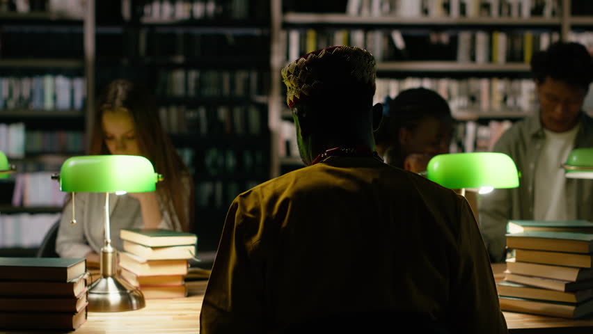 Back view of young adult with headphones studying at dark library table, framed by green lamps and stacks of books, Student consulting study materials near other peers.