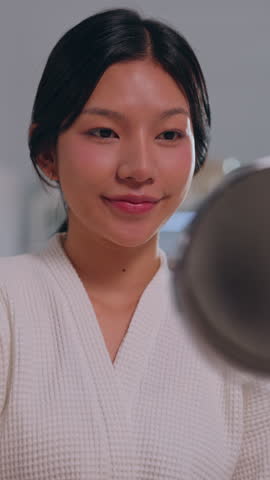 Happy Asian woman wearing white bathrobe looks into bathroom hand mirror during morning wellness skincare routine with cream, showing gentle smile on clean face for healthy beauty skin. Vertical